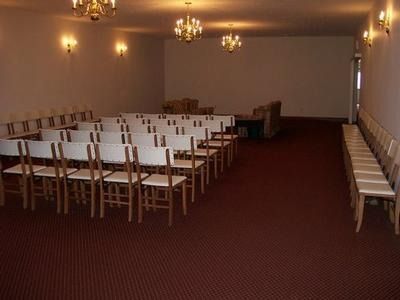 Rows of empty white chairs arranged in a large, dimly lit room with dark red carpeting and several chandeliers.