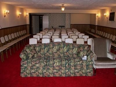 A furnished room with rows of chairs facing a floral sofa and a single armchair on a red carpet under warm wall lighting.