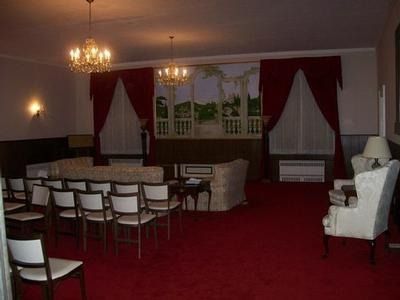 A dim formal event space with red carpet, arranged chairs facing a sofa, and large windows with red velvet drapes.