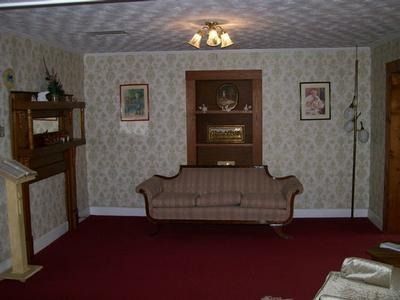A room with patterned wallpaper, red carpeting, a beige sofa, wood shelves, and a decorative fireplace.