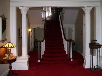 A grand foyer featuring red carpeting, a central staircase with white railings, and two large, white classical columns.