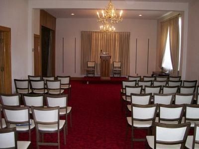 A room set up for a service, featuring rows of white chairs facing a wooden podium on a platform against a curtained wall.