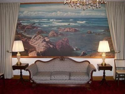 A patterned sofa sits centered beneath a large ocean landscape painting, flanked by two lamps on side tables and curtains.