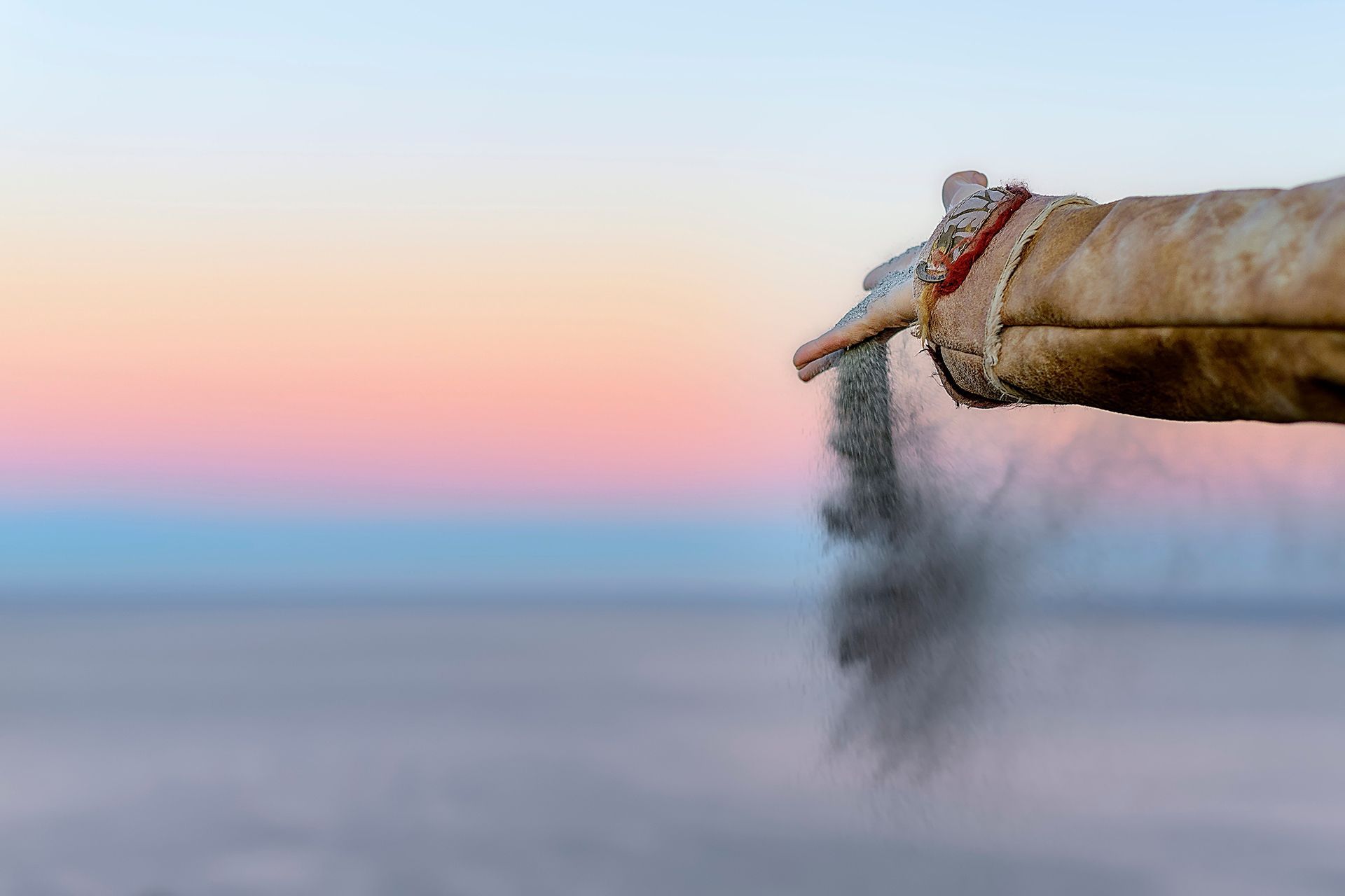 A gloved hand releases sand against a pastel-colored sky during twilight.