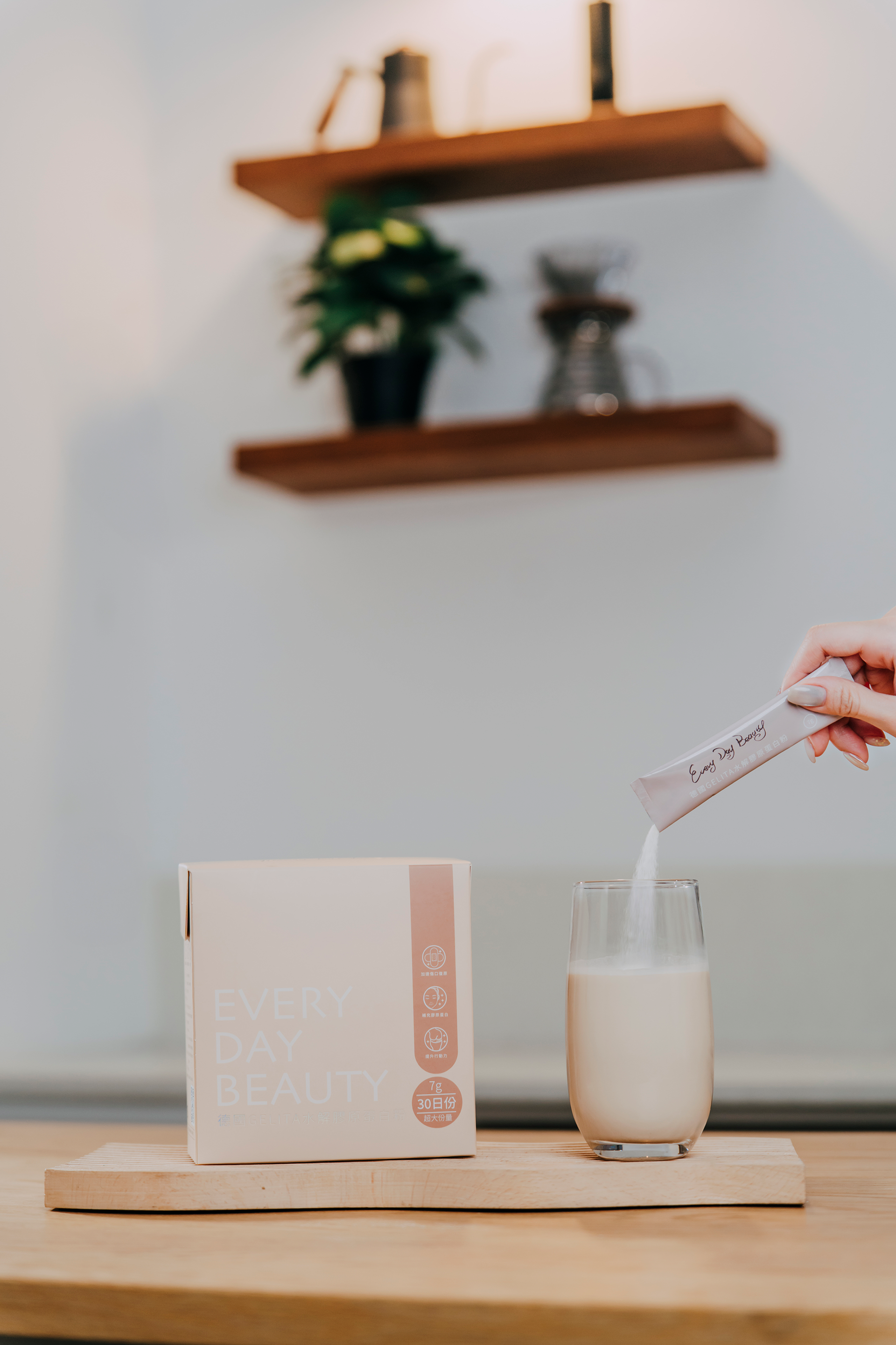 A person pouring powder from a packet into a glass of liquid, next to a box, on a wooden surface.