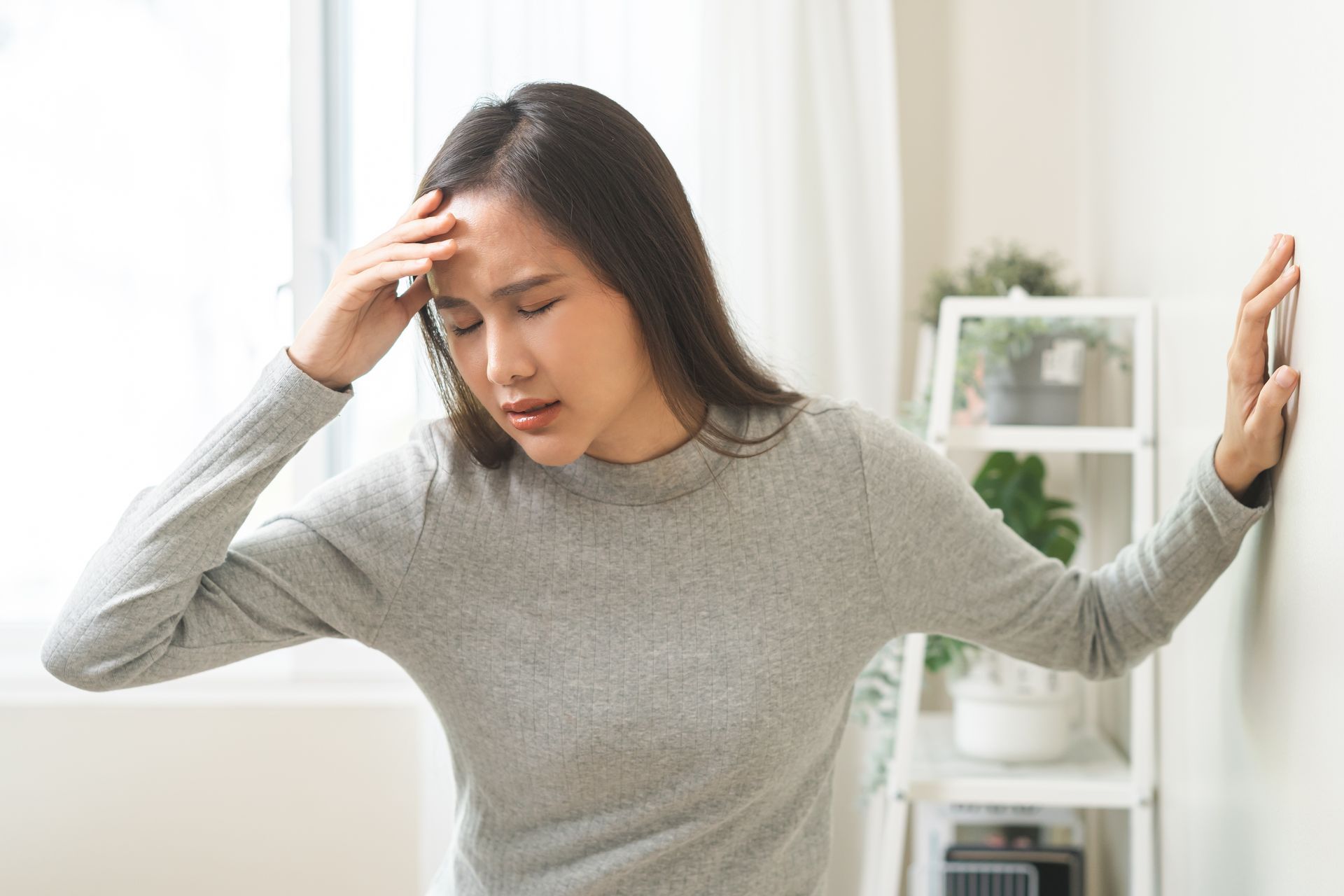 Young Asian woman in a grey sweatshirt holds the wall while suffering from dizziness due to TMD.