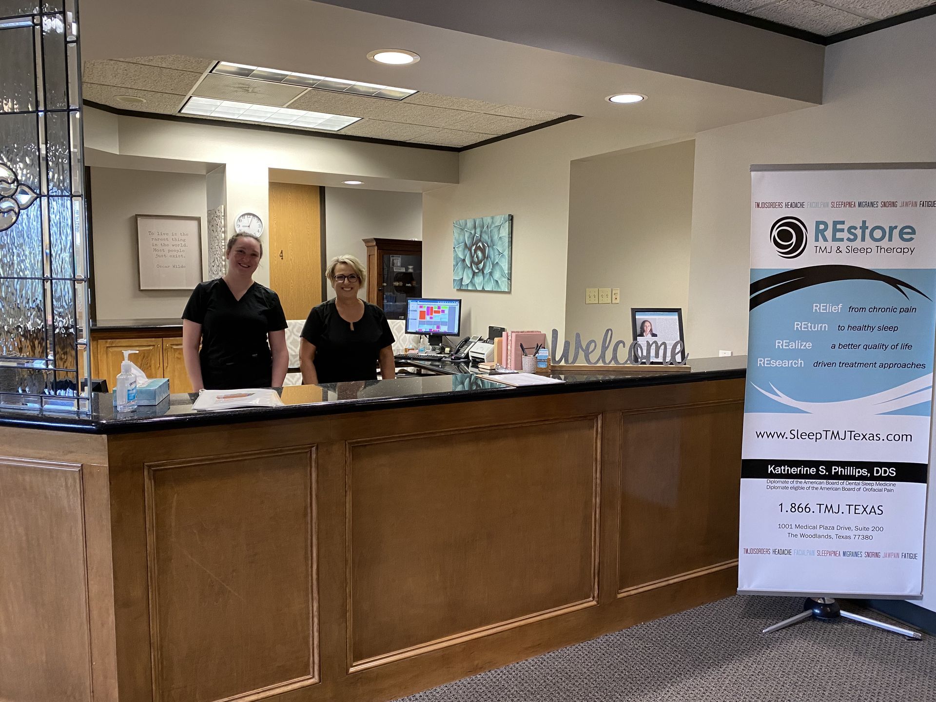 Two employees stand at the front desk and smile for the camera at the offices of Restore TMJ in Houston, Texas.