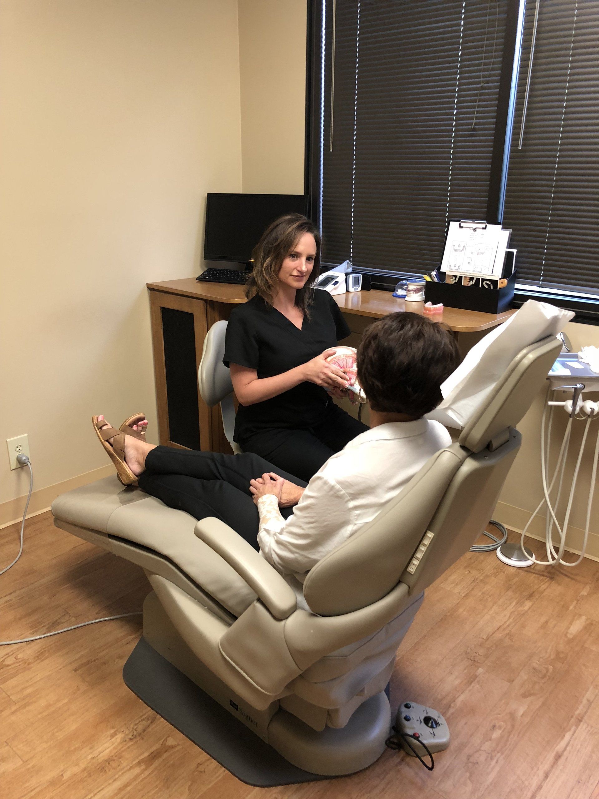 Female doctor in black scrubs speaks with female patient in white blouse sitting in patient's chair.