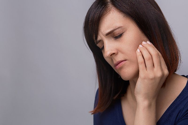 Woman with brown hair in blue blouse holds her jaw in pain from grinding her teeth.