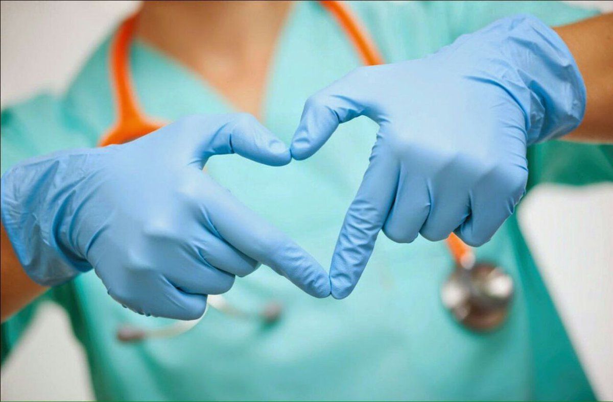 Close up on medical personnel in scrubs making the shape of a heart with their fingers in blue surgical gloves.