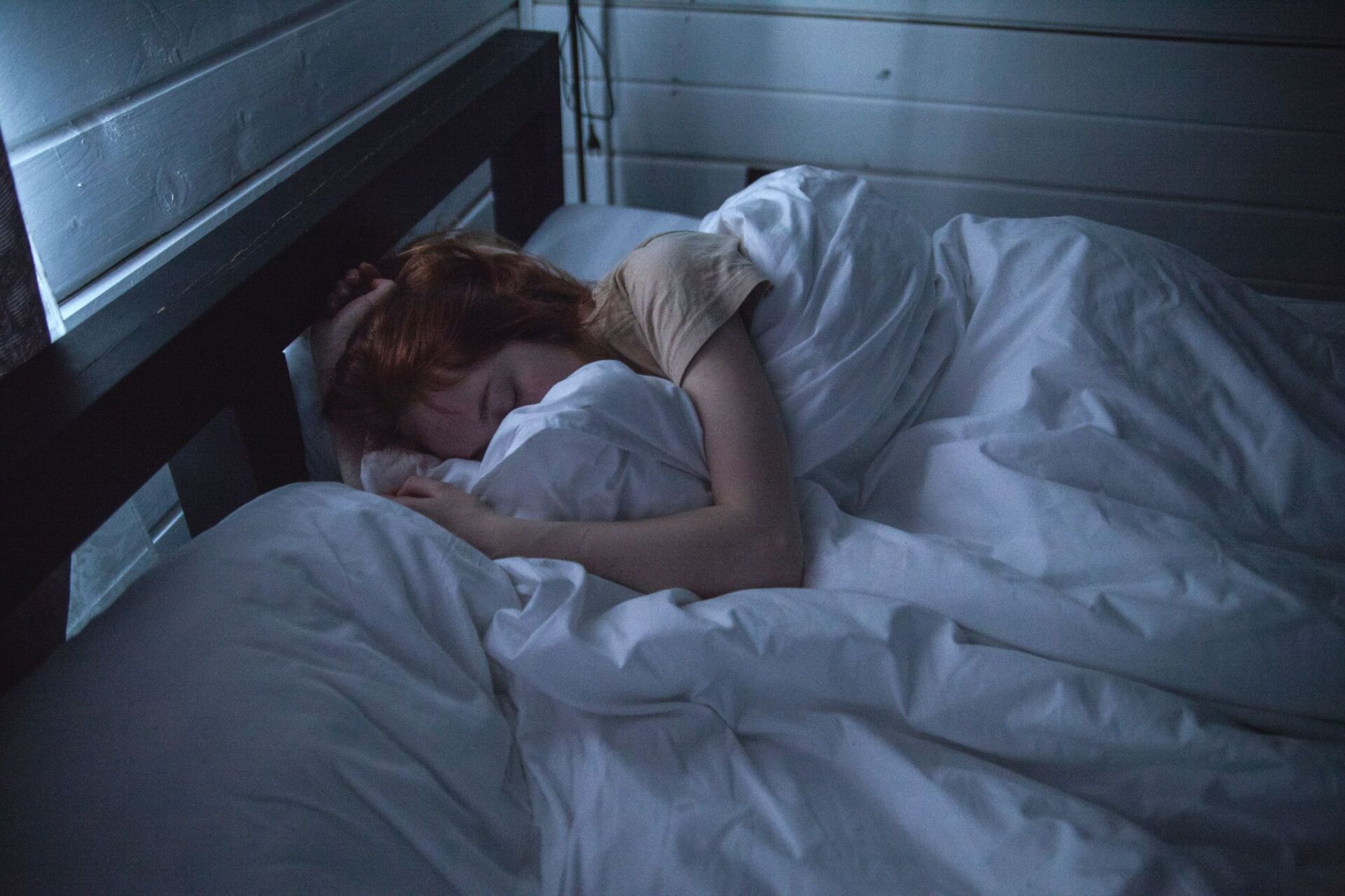 Young redheaded woman curled up under a white comforter while sleeping in bed.