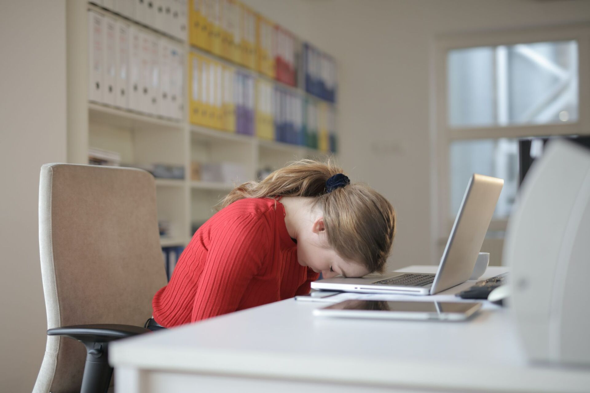 Woman in red shirt sleeping at computer desk with her head rested on a silver laptop.