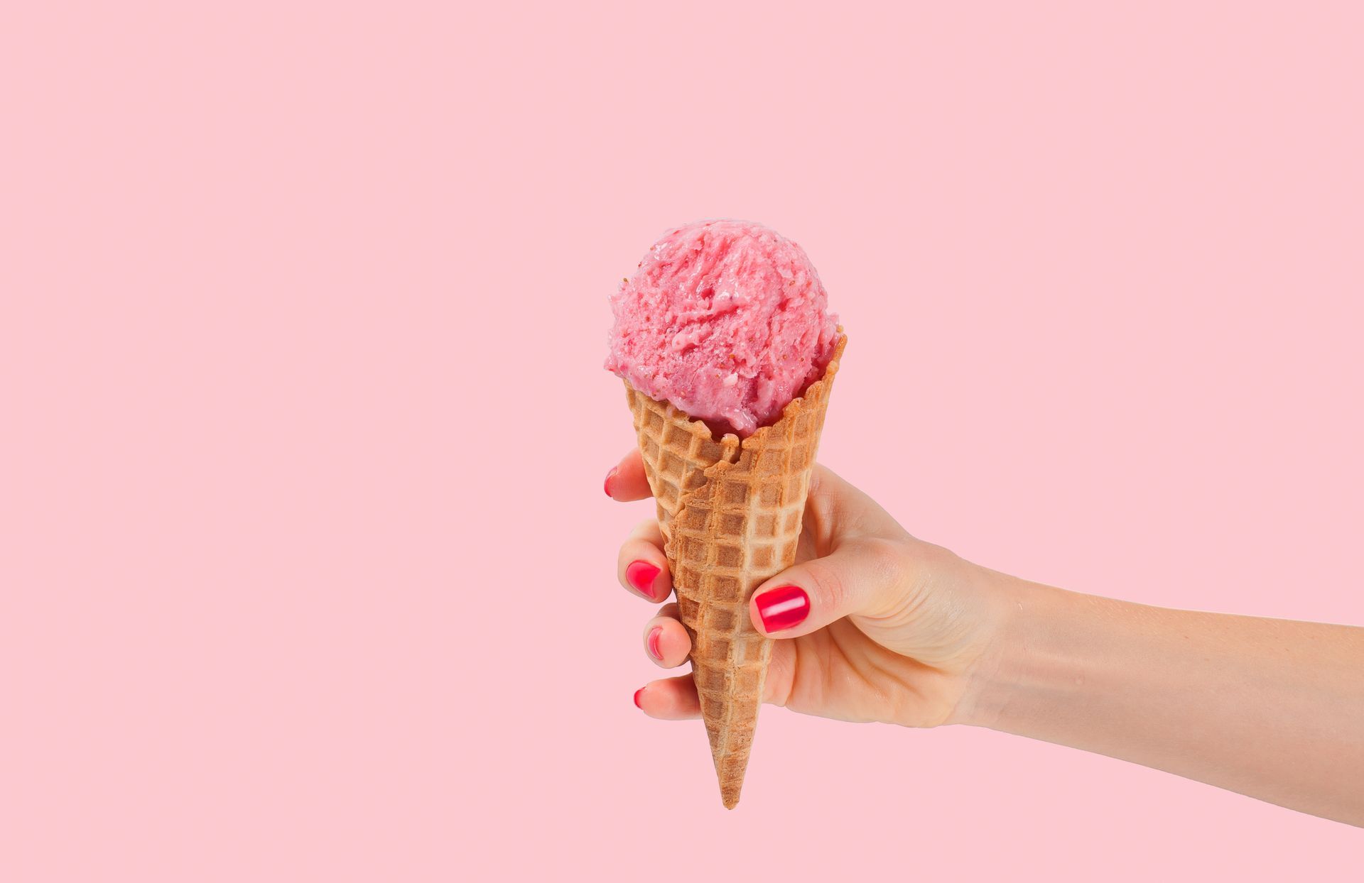 Hand holding a waffle cone with a scoop of pink ice cream against a pink background.