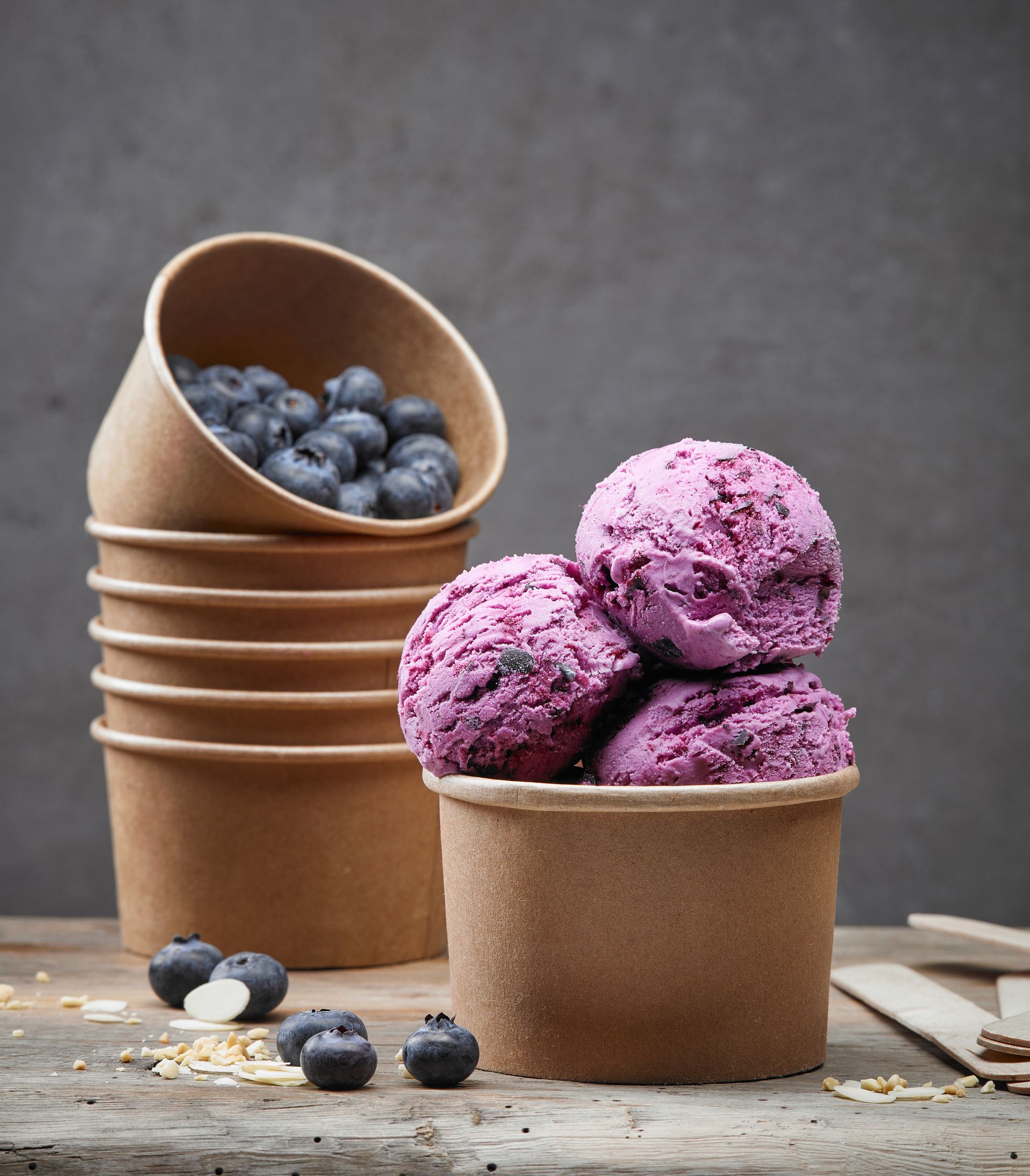 Blueberry ice cream scoops in a brown paper cup, with more cups and blueberries nearby.