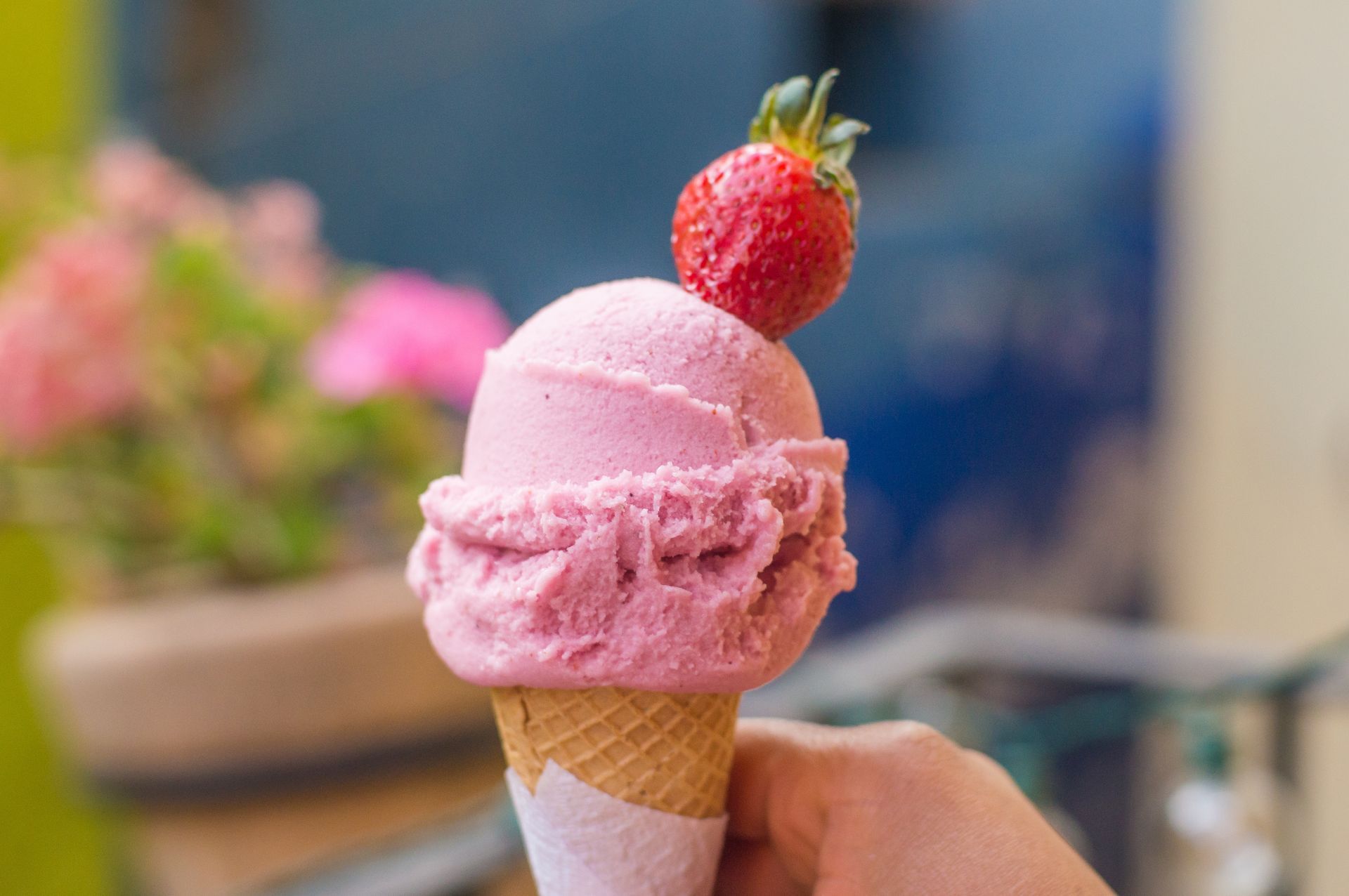 Hand holding a pink ice cream cone topped with a strawberry, with blurred flowers in the background.