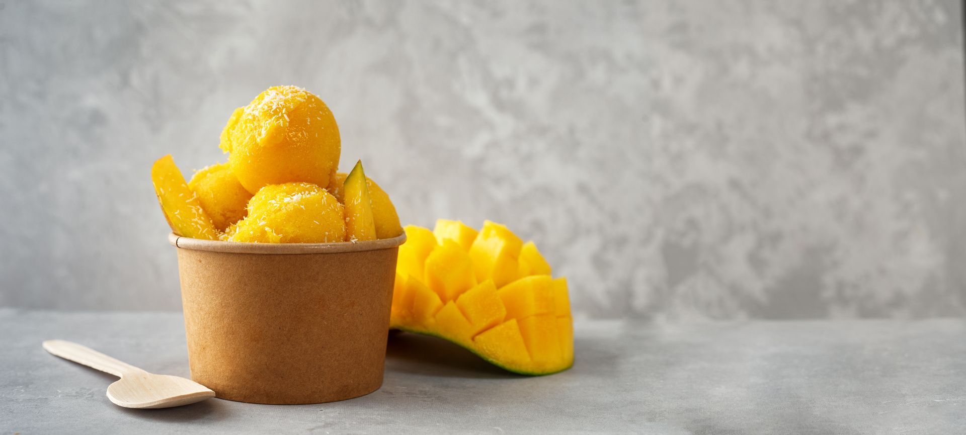 Mango sorbet scoops in a brown cup with a wooden spoon and mango slices on a gray surface.