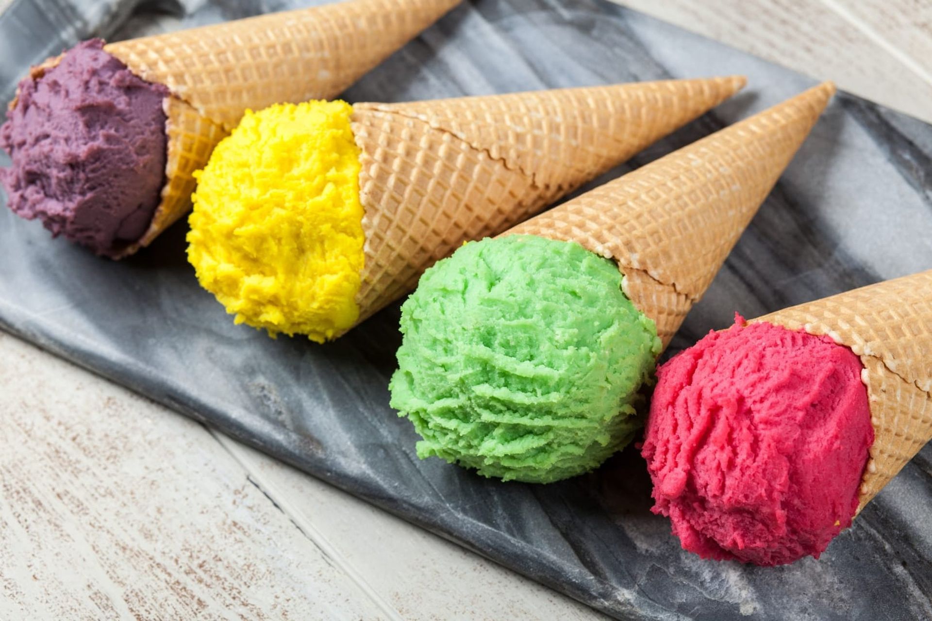 Four ice cream cones with colorful scoops: purple, yellow, green, and pink, on a marble tray.