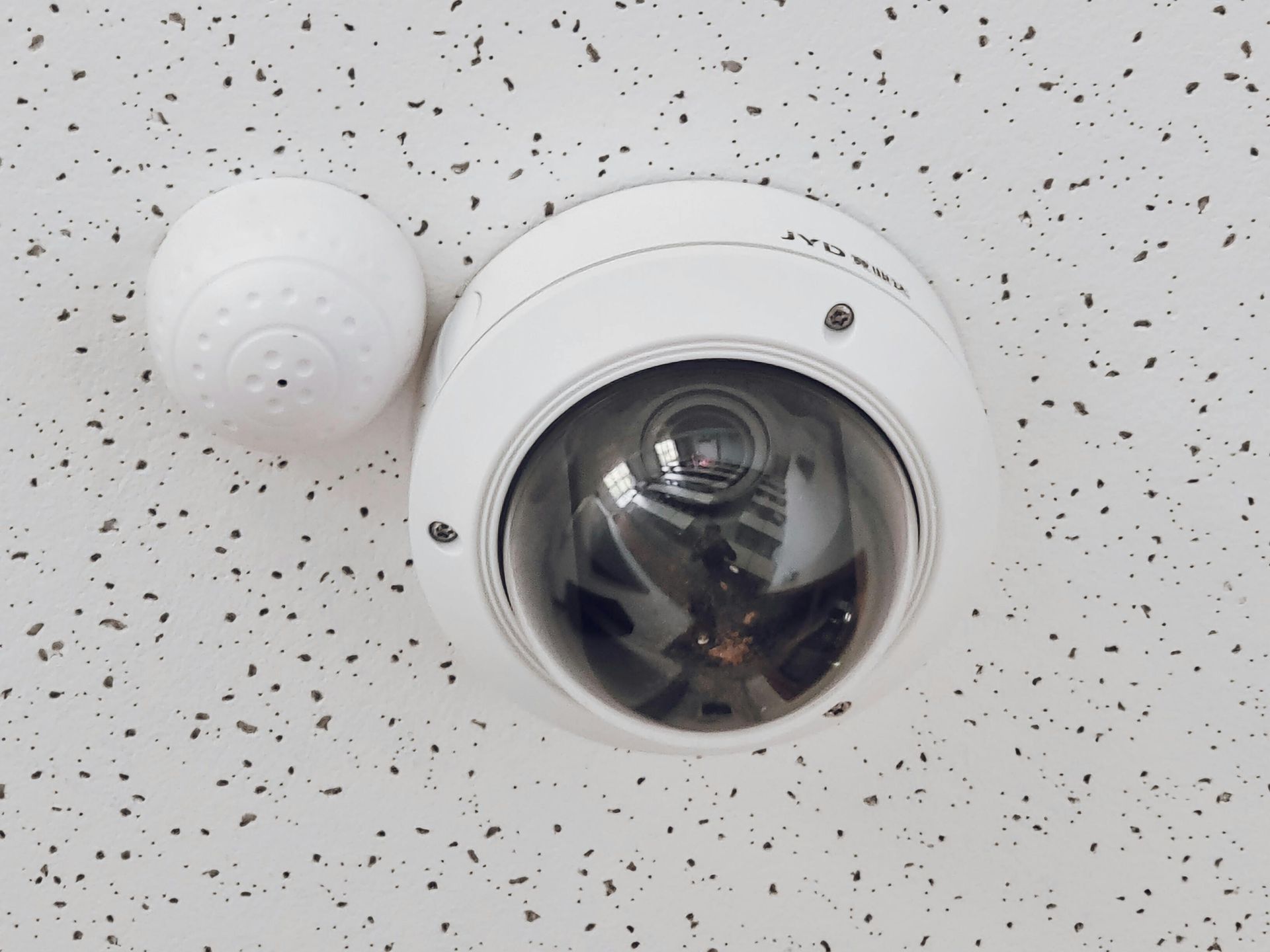 A security camera and a small, round device are mounted on a white ceiling with dark speckles.