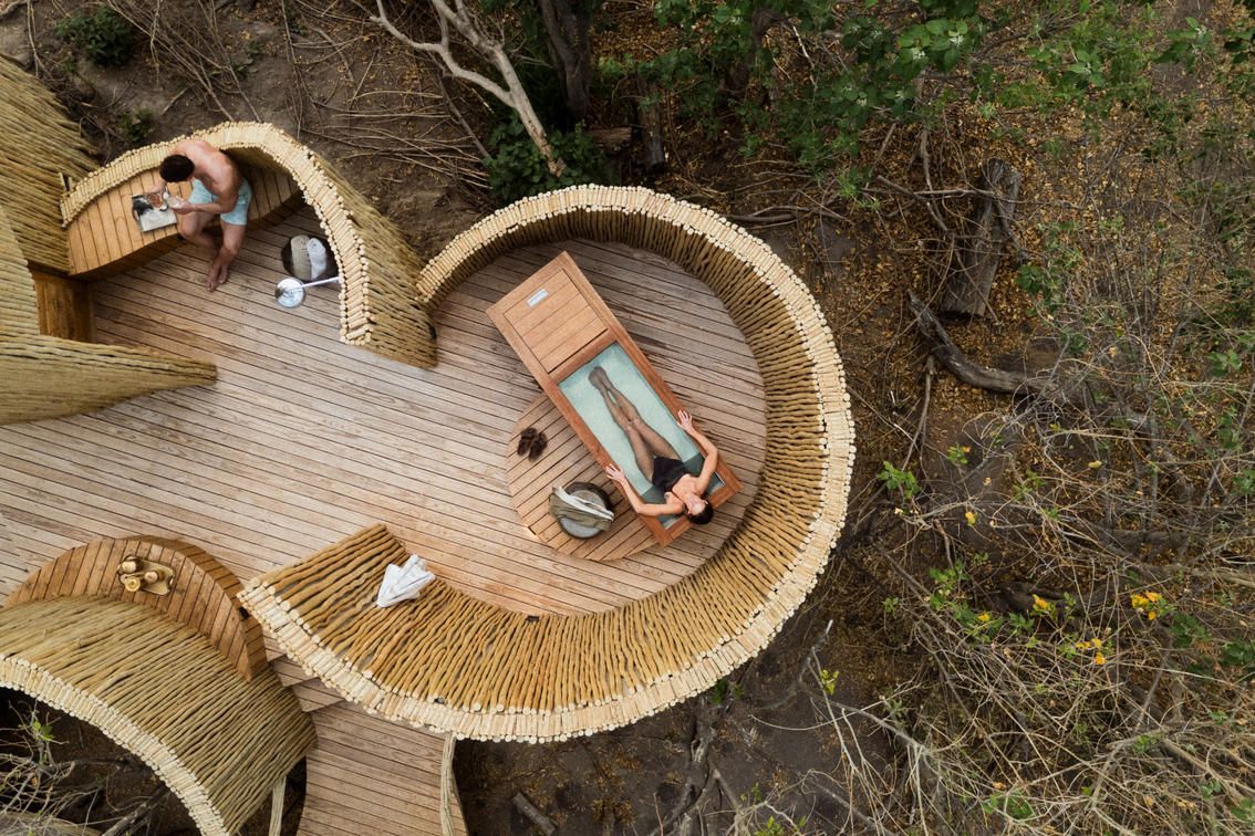 An aerial view of a person relaxing on a wooden lounger inside a circular, straw-walled structure in a forest.