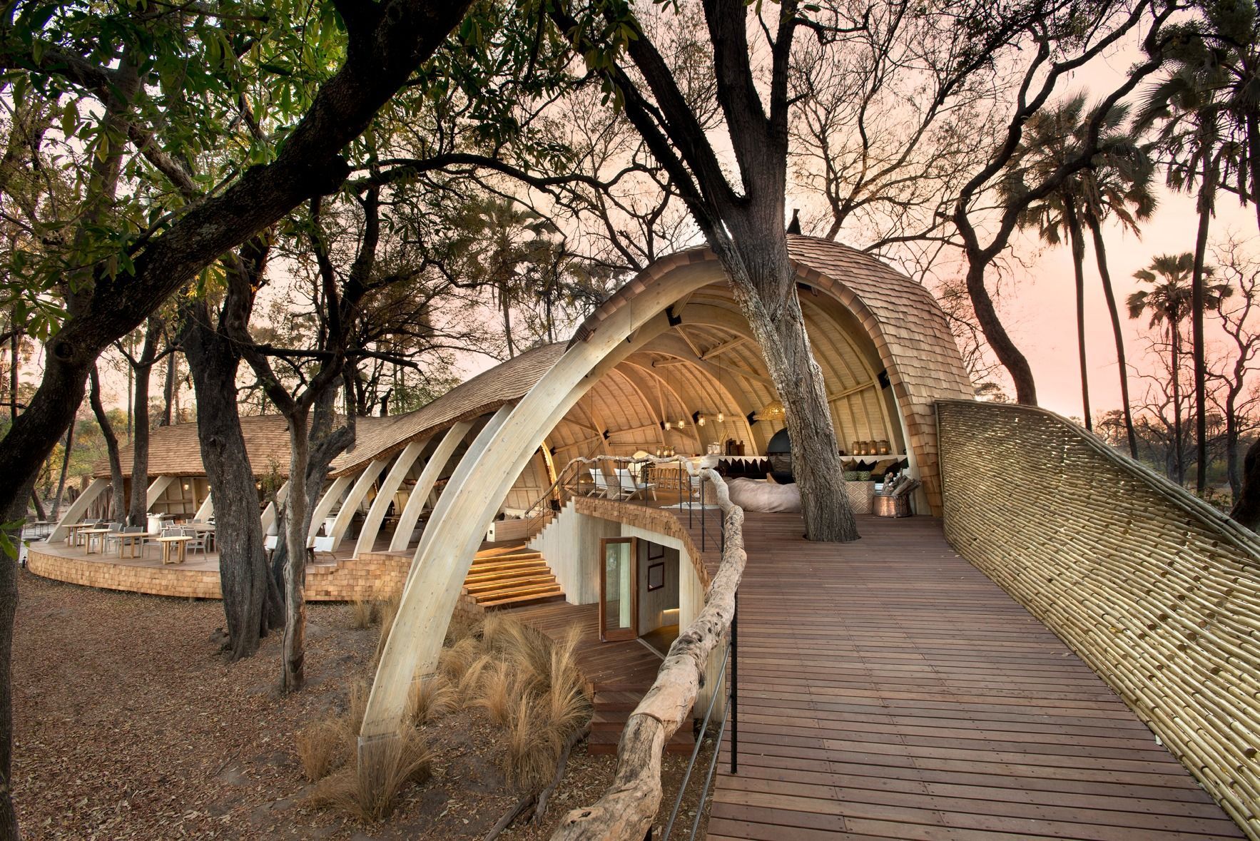 A wooden walkway leads to a modern, arched, thatched-roof structure nestled among trees in a safari-like setting.