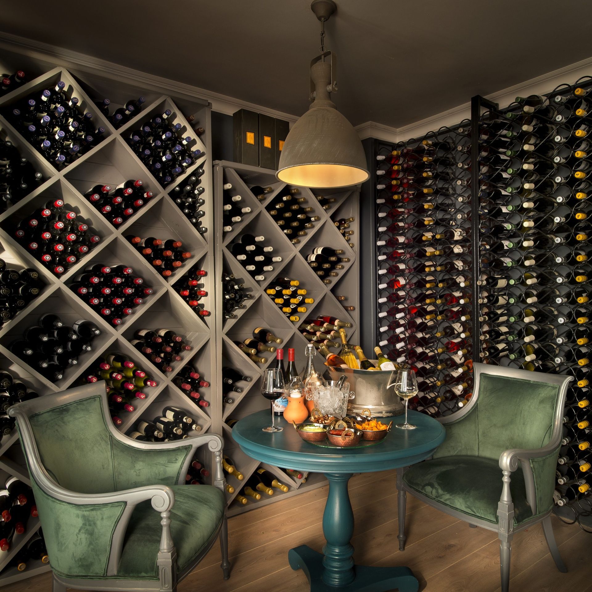 Two velvet green chairs and a small table set in a dimly lit, cozy wine cellar with extensive wall-to-wall bottle racks.