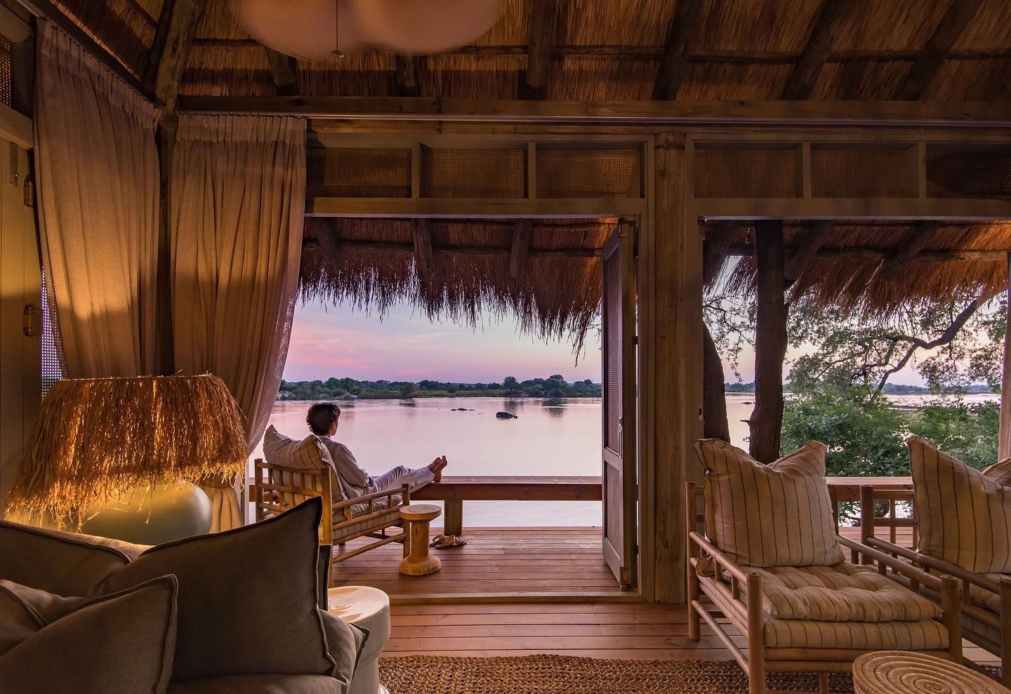 A person relaxes on a wooden deck overlooking a calm river at sunset, framed by a rustic open-air lodge interior.