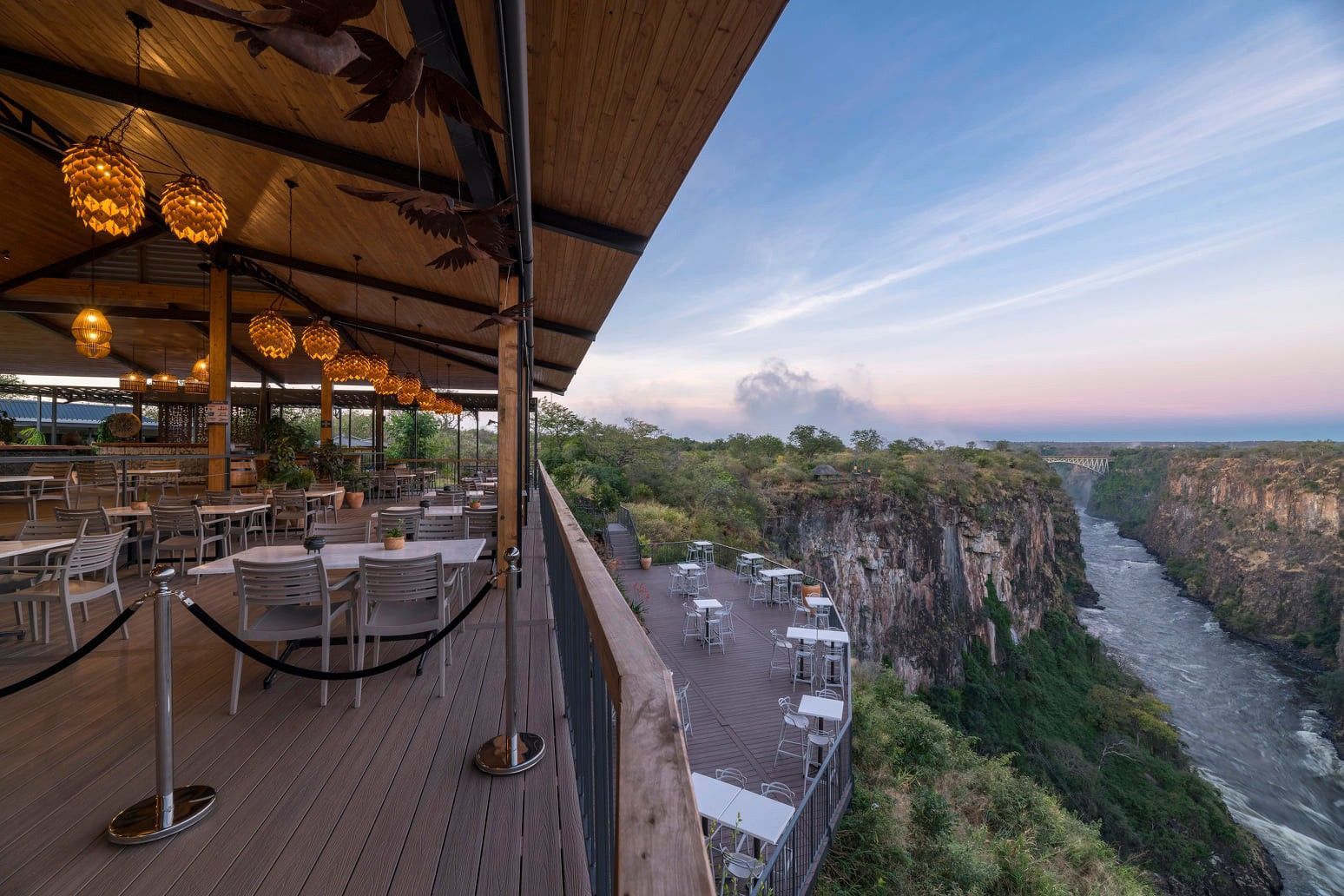 Outdoor restaurant terrace overlooking a river canyon at sunset, with dining tables set up on both upper and lower levels.
