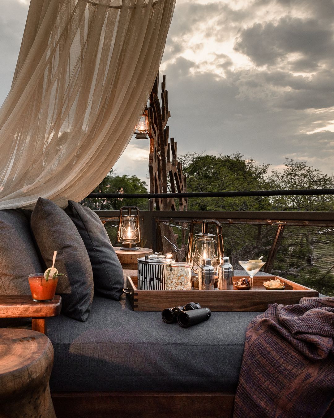 A cozy outdoor seating area with grey cushions, a wooden tray of drinks, lanterns, and binoculars under a canopy.