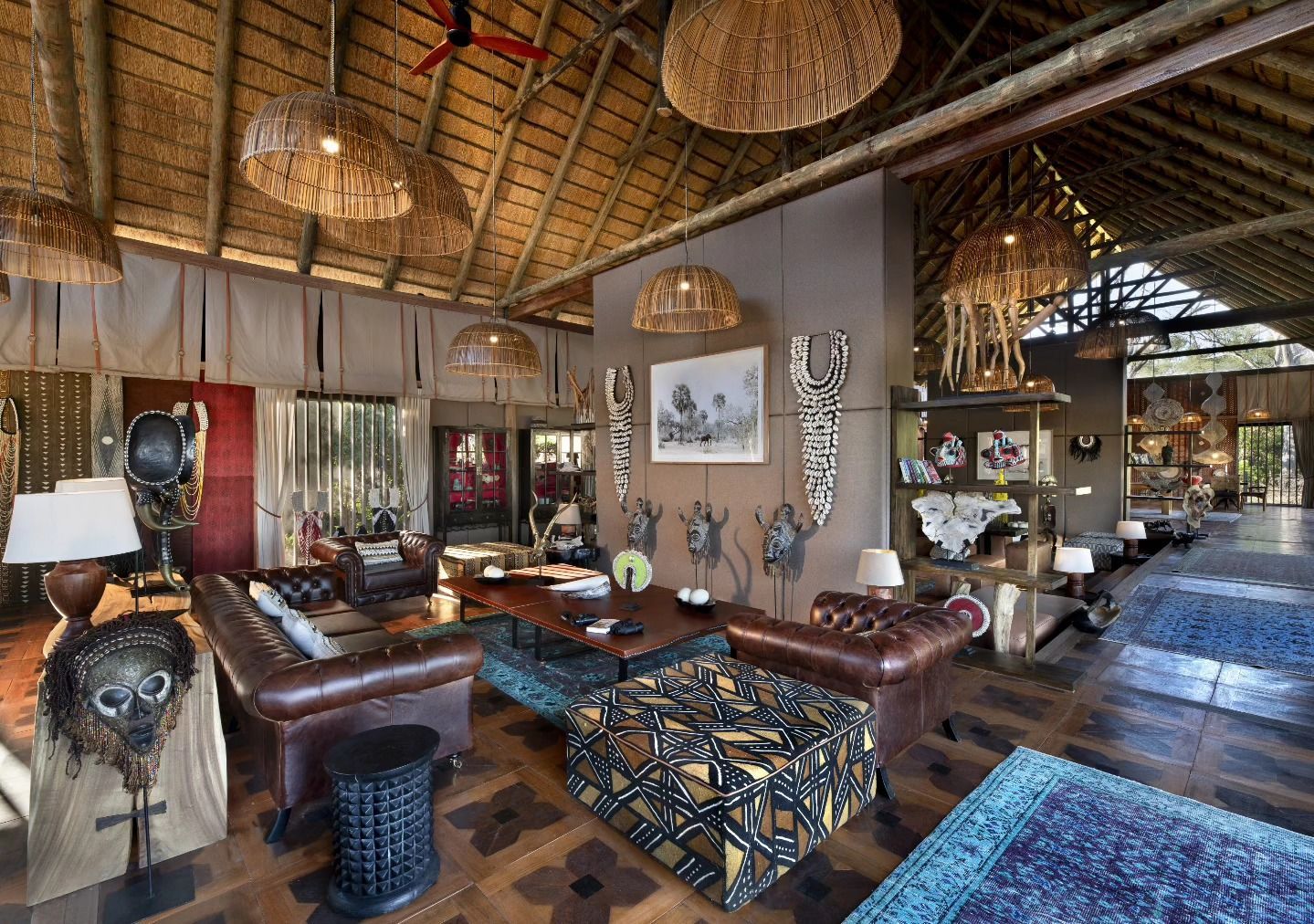 A safari-style lodge living area with a high thatched roof, leather sofas, patterned ottomans, and African art decor.