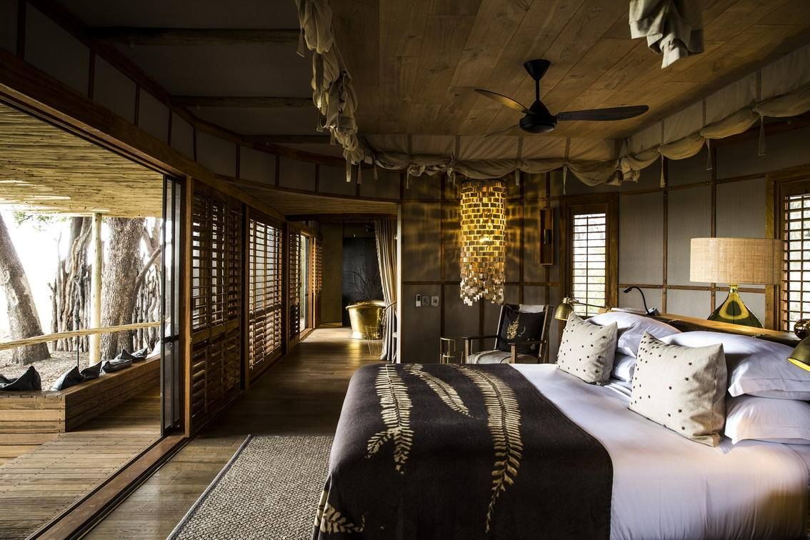 A luxurious, rustic bedroom suite with a large bed, wooden walls, a ceiling fan, and views of a tree-filled outdoor deck.