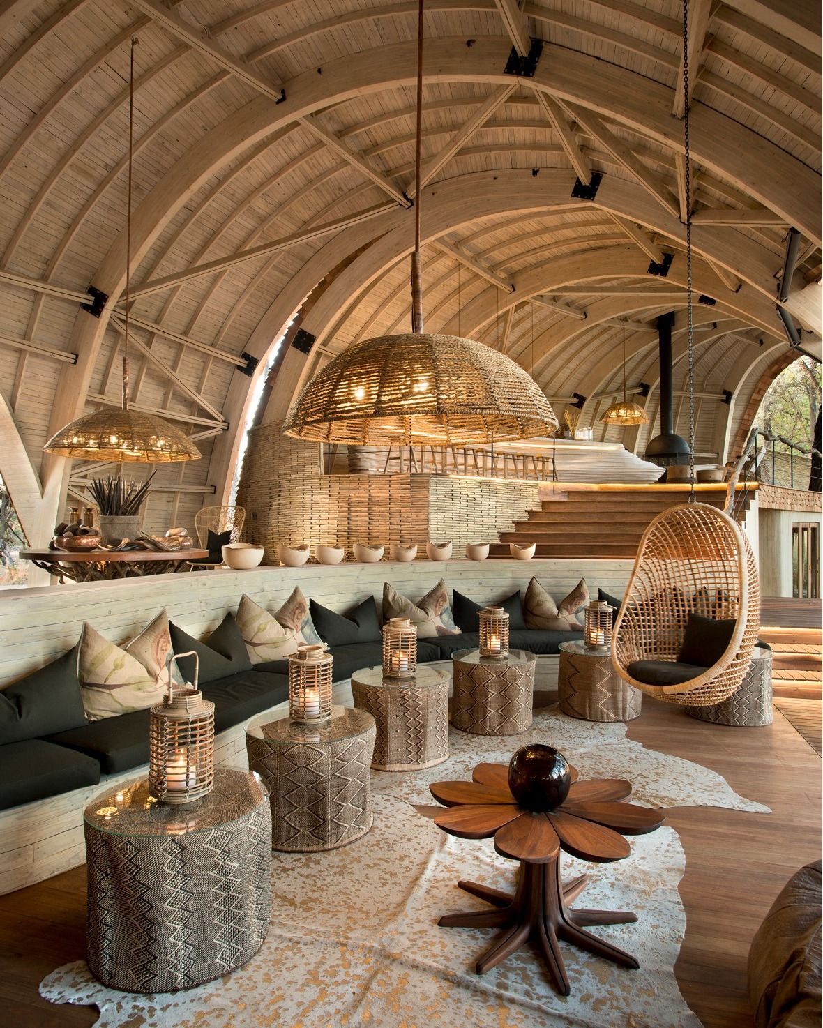 A warm, rustic lounge featuring a curved wooden ceiling, low seating, lantern-style lamps, and a hanging wicker chair.