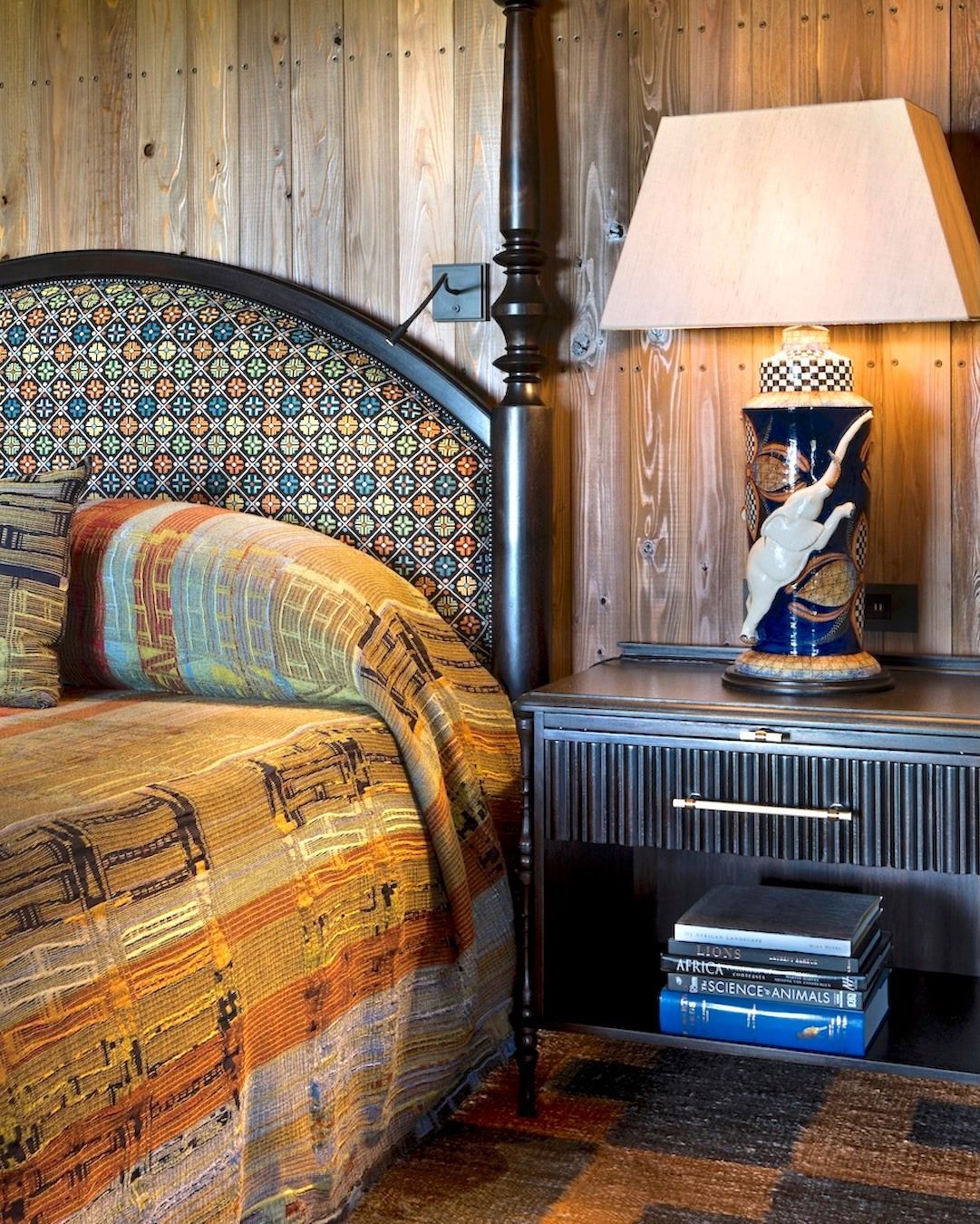 A rustic bedroom corner featuring a patterned headboard, a tribal-print quilt, and a blue ceramic lamp on a nightstand.