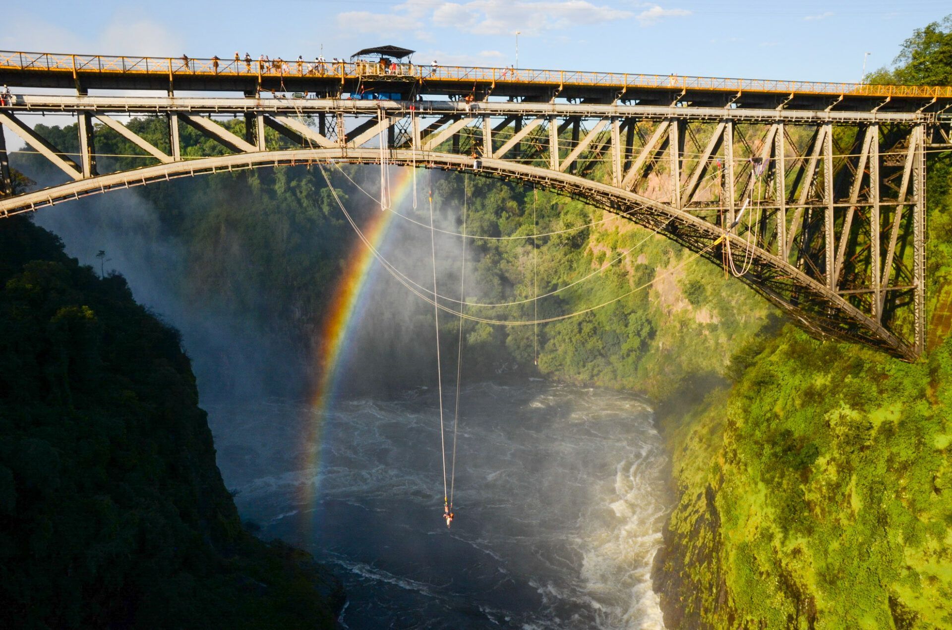 A person bungee jumps from the Victoria Falls Bridge, with a bright rainbow arching through the mist below.