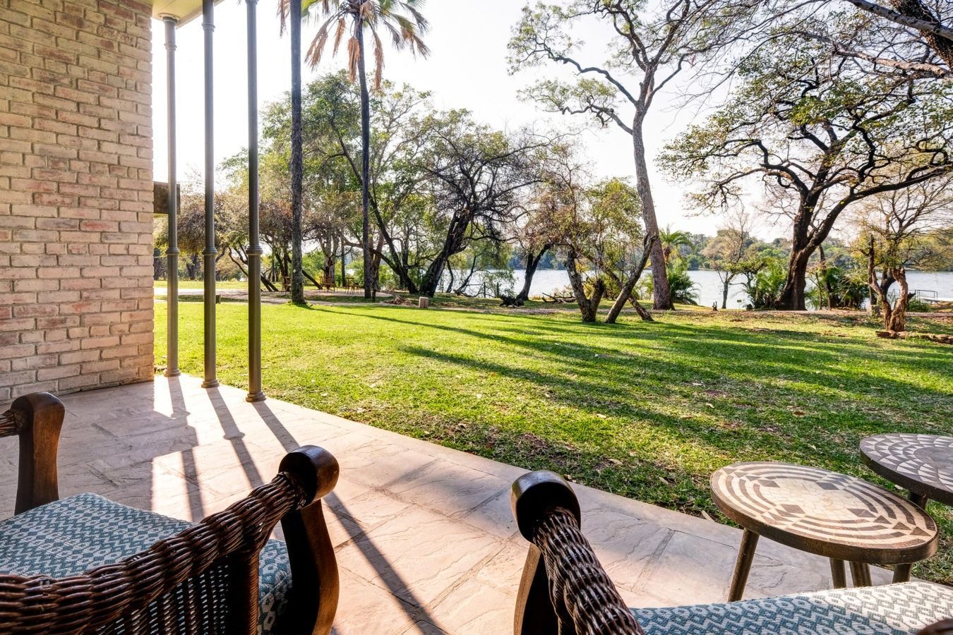 Wicker armchairs on a paved patio looking out onto a grassy lawn with trees and a river in the background.