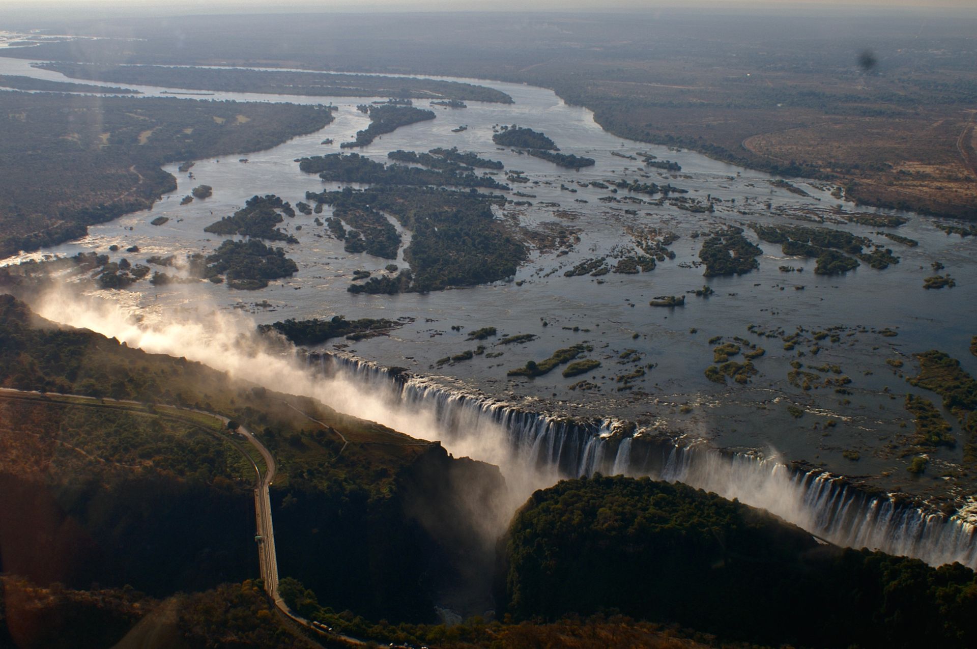 Aerial view of Victoria Falls, with the Zambezi River flowing toward the cliff edge and a bridge spanning the gorge.