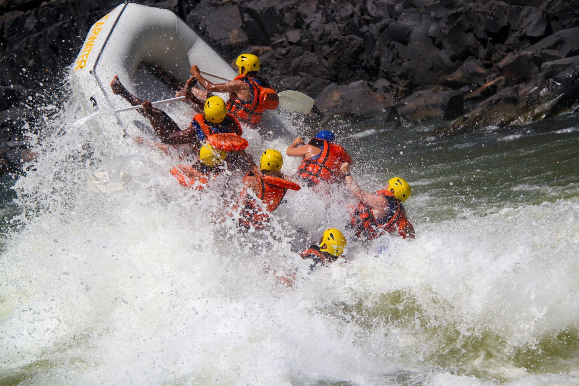A whitewater raft tipped vertically on a wave with several rafters in life jackets and yellow helmets falling into water.