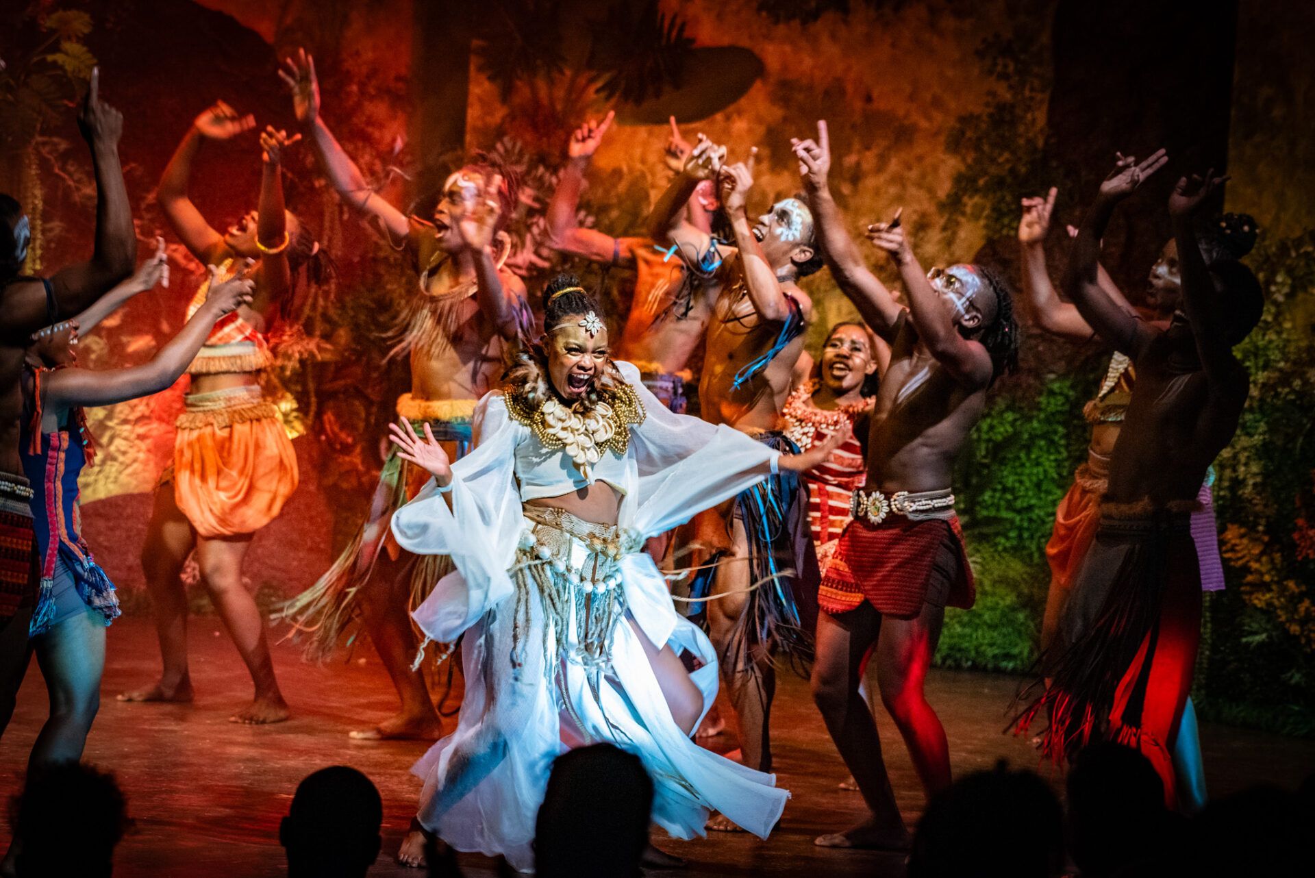 A group of performers in vibrant, tribal-style costumes dancing together on a stage under warm lighting.
