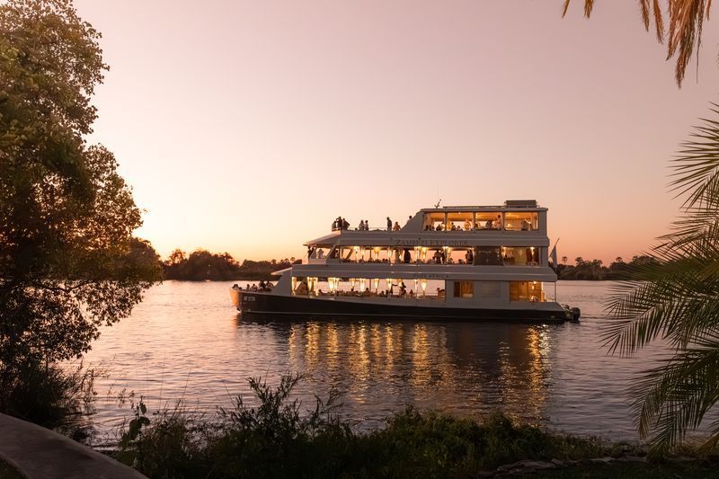 A multi-level riverboat cruises on a calm waterway during a pink sunset, framed by trees on both sides.