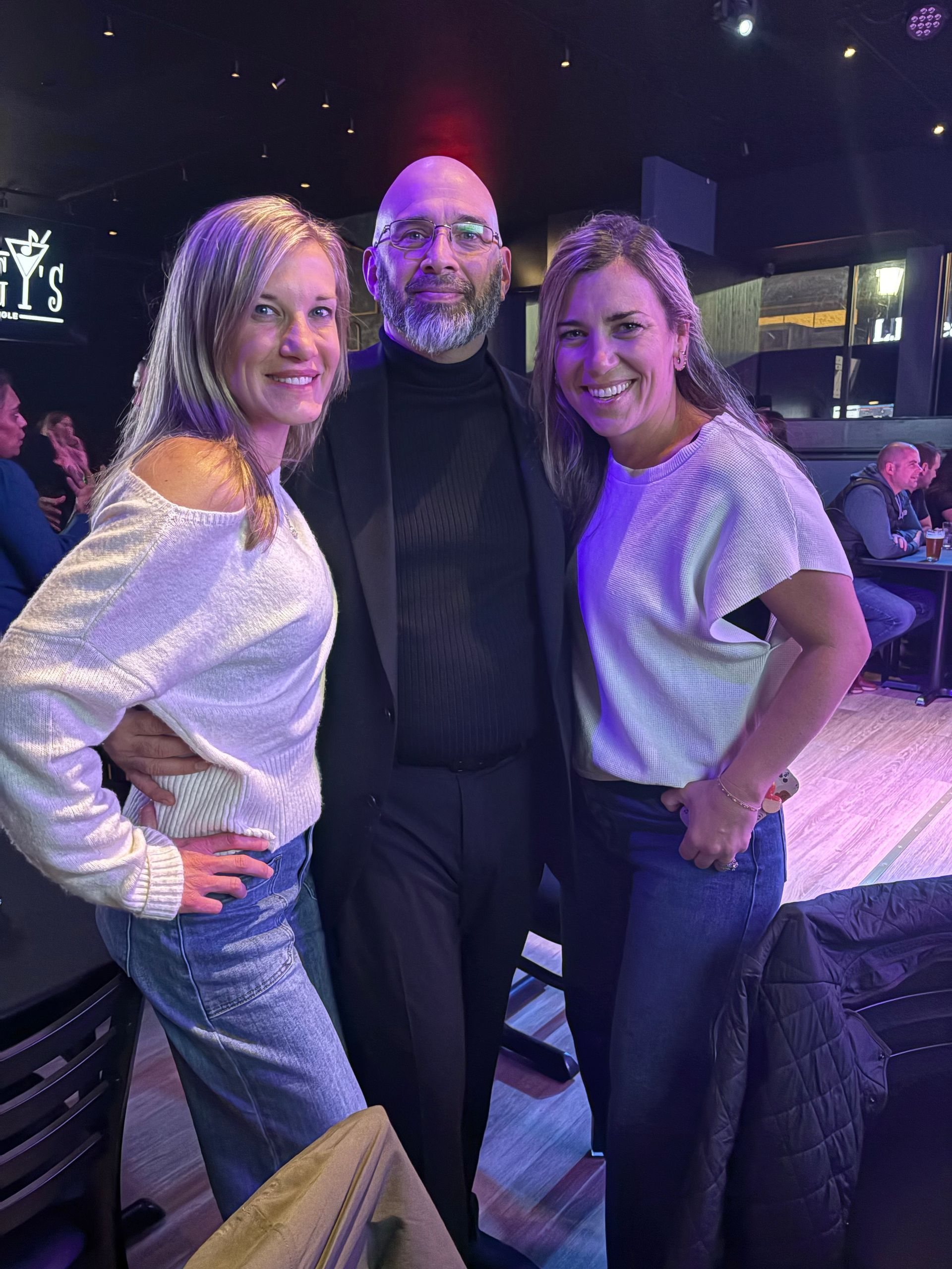 Three people posing in a dimly lit bar. Two women flank a man. All are smiling.