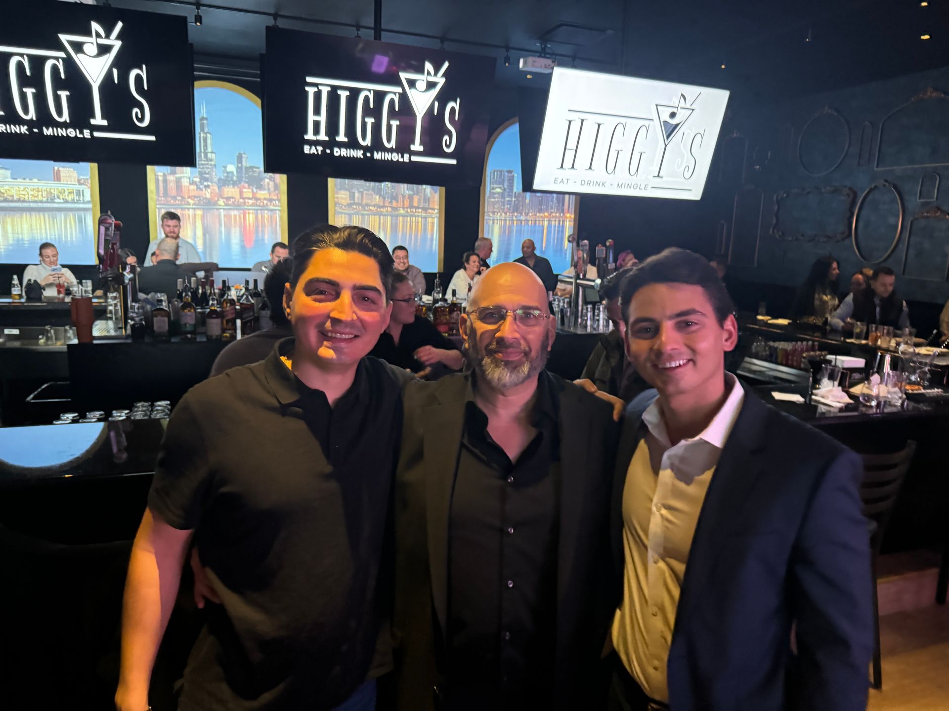 Three men smiling, posing at Higgy's restaurant, with bar and tables visible.