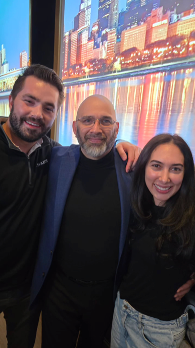 Three people posing for a photo. A man with a beard is between a man and a woman in front of a city scene backdrop.