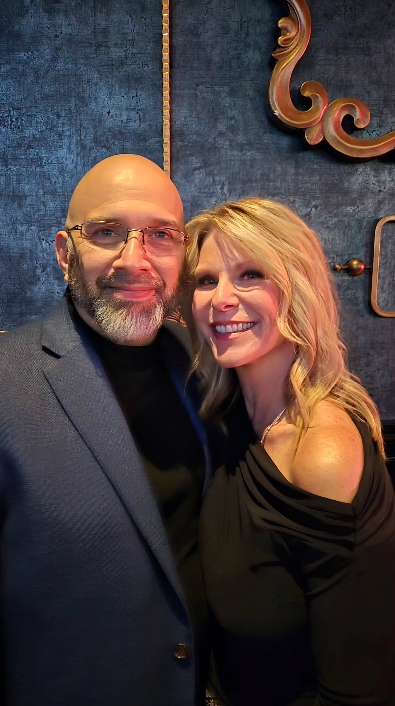 Man and woman smiling, indoors. Man in glasses and blazer, woman in black top, against a blue and gold wall.