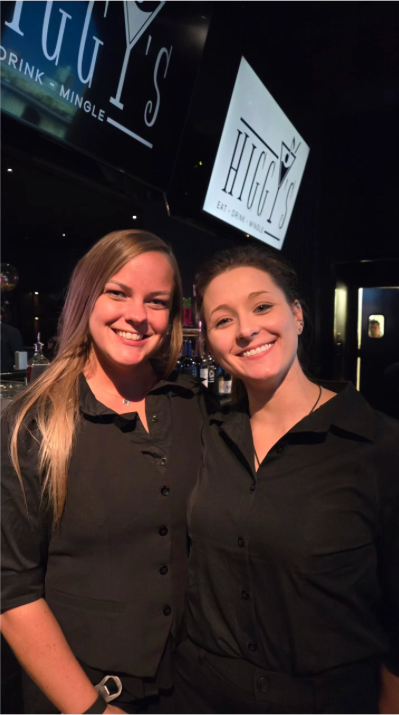 Two smiling people in black shirts pose at a bar under a sign that reads 