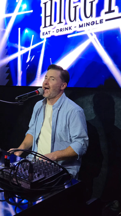 Man singing at a venue, blue lighting in background, microphone, mixing board, stage.