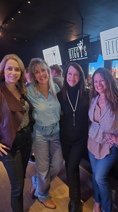 Four women pose indoors. Two are smiling and holding each other. The setting appears to be a bar or restaurant.