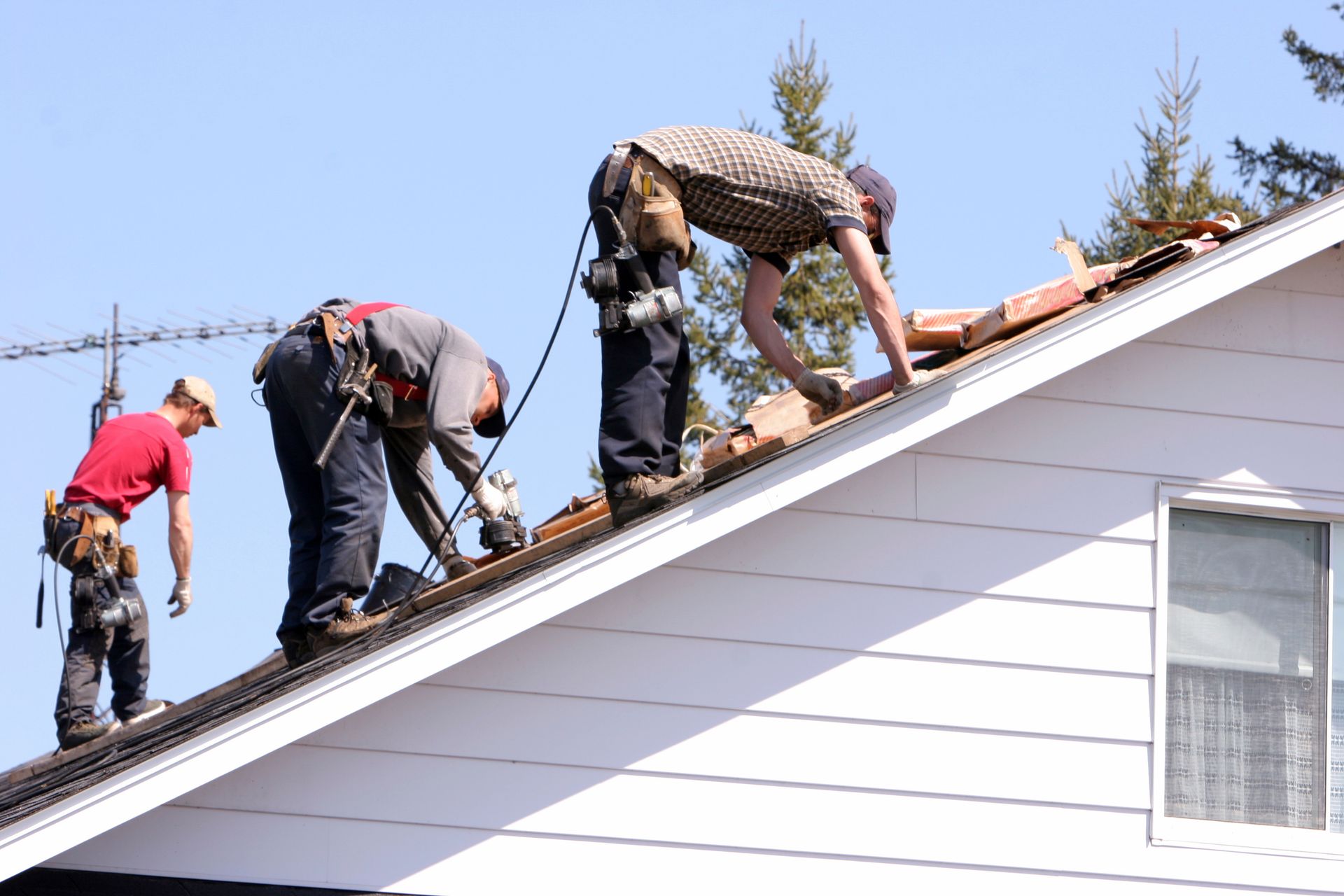 Three roofers working on a white-sided house roof under a sunny sky.