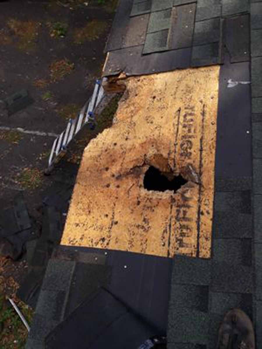 Damaged roof showing a large hole in the plywood with missing shingles, daylight.