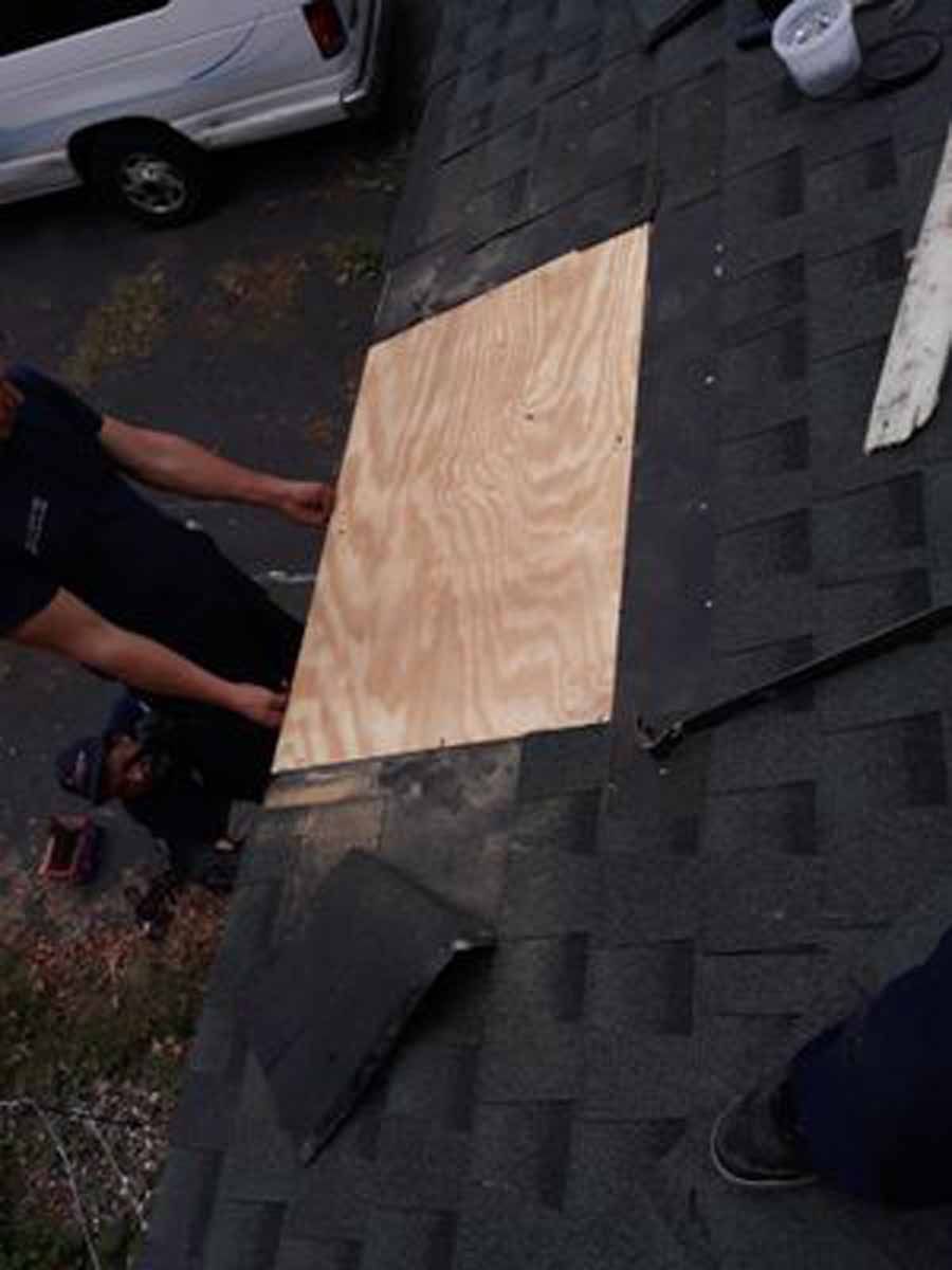 Two people on a roof placing a plywood patch over a missing section of shingles.