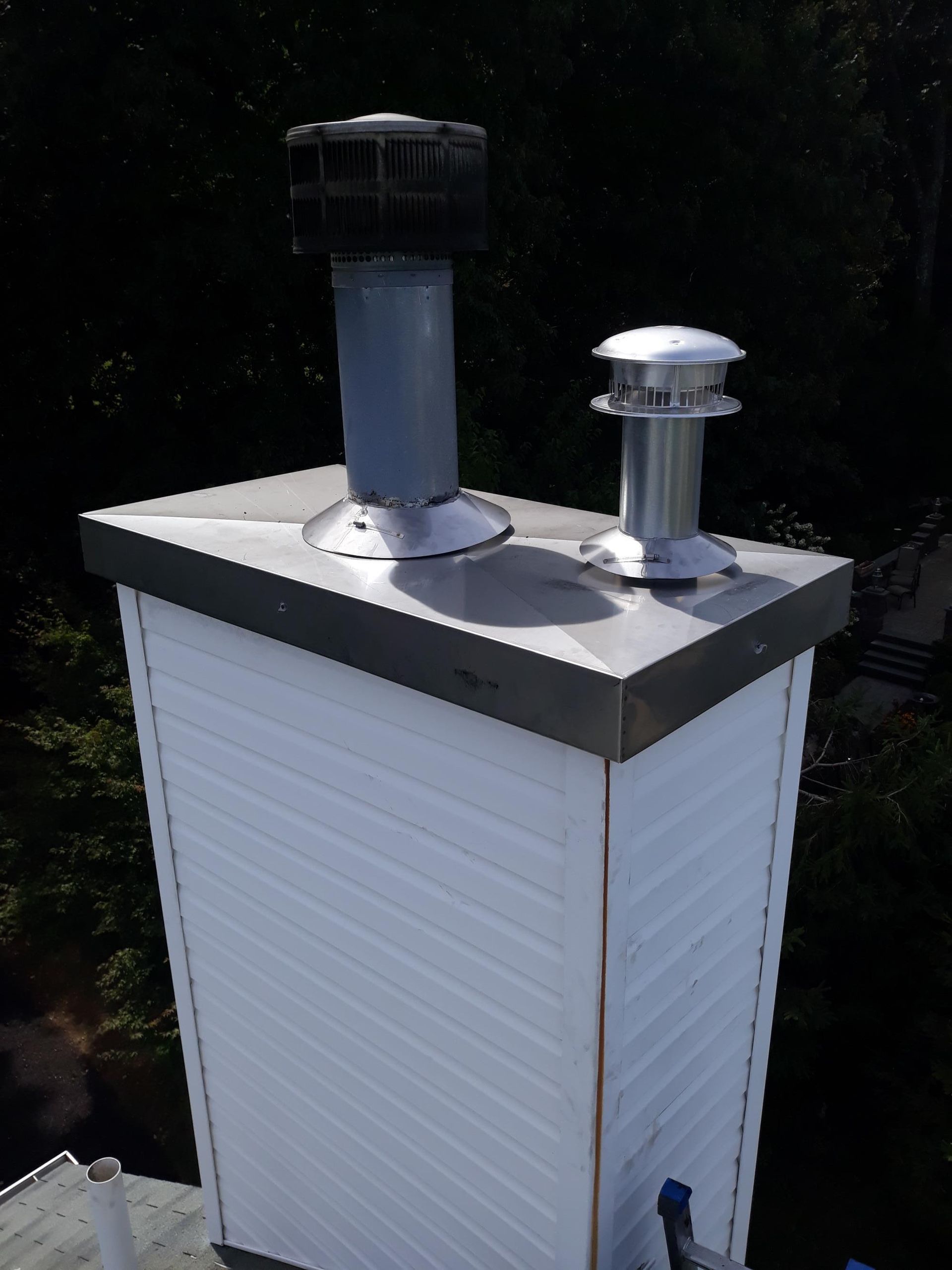 Chimney with two metal flue caps, white siding, and dark trim, against a dark green backdrop.