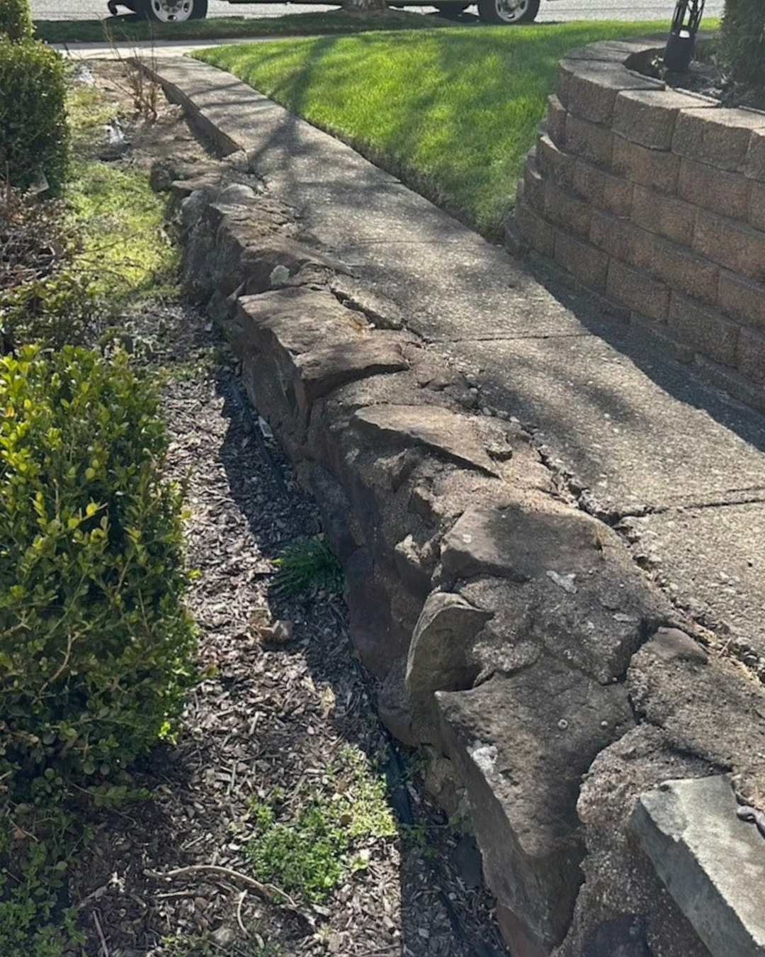 Cracked stone retaining wall along a sidewalk, with grass and shrubbery on either side.